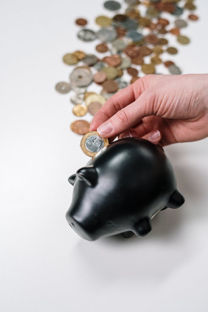 A hand putting a coin into a black piggy bank with scattered coins on a white background.