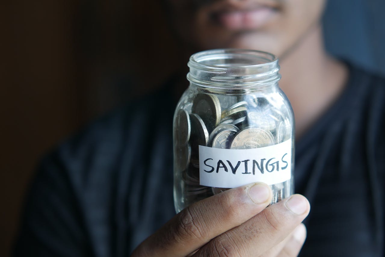 A close-up image of a persons hand holding a jar full of coins labeled Savings.